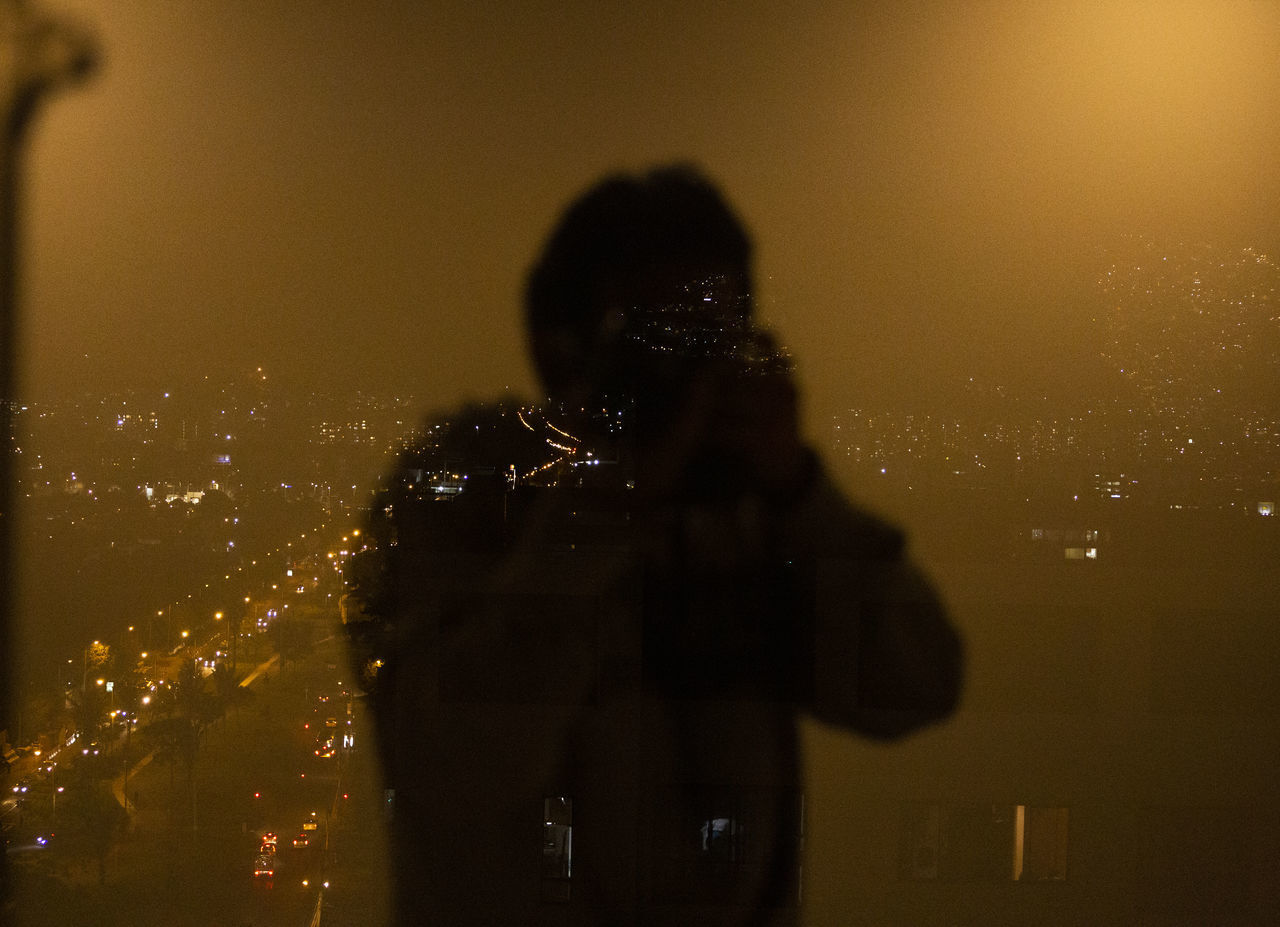 SHADOW OF MAN PHOTOGRAPHING ON ILLUMINATED CITYSCAPE AT NIGHT