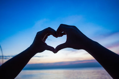 Close-up of hand making heart shape against sky during sunset