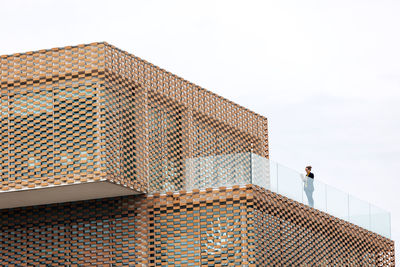 From below of female in stylish outfit standing on balcony of modern building with geometric elements on windows while using tablet near glass railings under bright sky