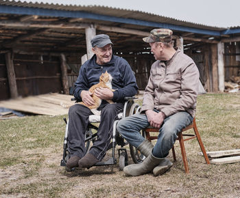 A happy senior man in a wheelchair in the yard , smiling, holding a cute cat, talking to his friend.