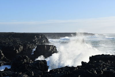 Waves splashing on rocks at shore against sky