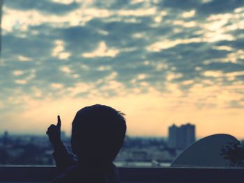 Rear view of boy pointing while standing on building terrace against cloudy sky during sunset
