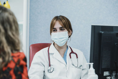 A young and good looking female doctor with face mask is talking to a patient in her office. 