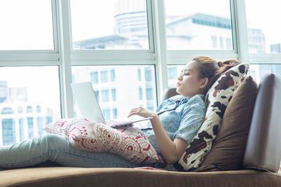 Young woman using mobile phone while sitting on window