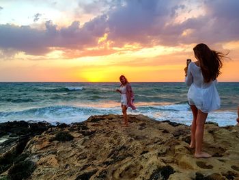 Woman standing on rock at beach against sky during sunset