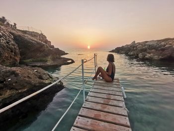 Woman sitting on rock by sea against sky during sunset