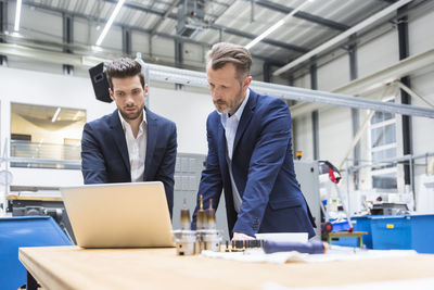 Two men at table in factory using laptop