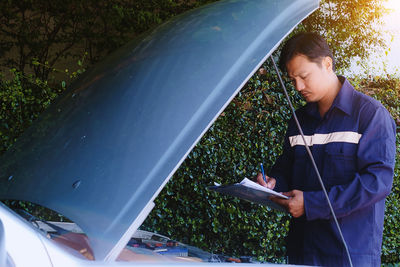 Mechanic writing in notepad by car against plants