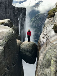 Woman standing on kjeragbolten rock with waterfall, fjord, mist, clouds, and mountains in background 