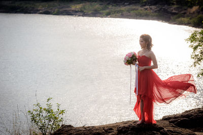 Full length of bride wearing red wedding dress holding bouquet while standing on rock by lake