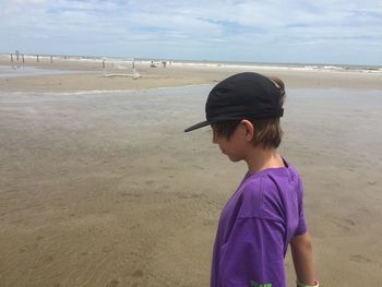 Side view of teenage boy wearing cap standing at beach against sky during sunny day