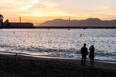 Rear view of silhouette friends standing at beach during sunset