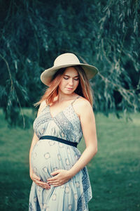 Smiling pregnant woman touching abdomen while standing on grass