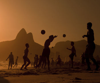 Silhouette friends playing soccer at beach against orange sky during sunset