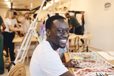 Portrait of happy male artist sitting with paintings at table in art class