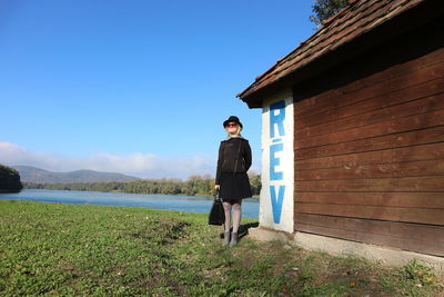 Full length of woman by log cabin at riverbank against blue sky