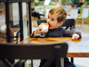 Toddler boy in casual clothes eating a chocolate bun in a food court there is chocolate on his face