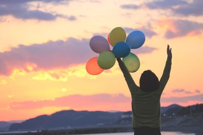 Girl standing with arms raised against sunset sky and holding balloons up