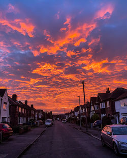 Road by buildings against sky during sunset