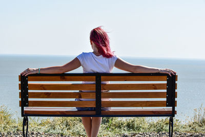 Rear view of woman sitting on bench by sea against clear sky