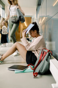 Female teenager with book covering face while sitting in school corridor