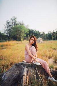 Portrait of young woman sitting on tree stump against clear sky