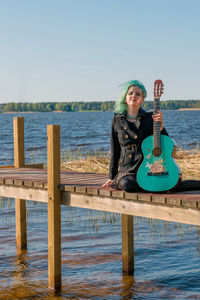 Hipster young woman with turquoise guitar sitting on pier over lake