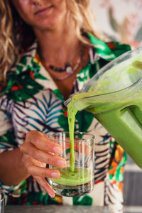 Female pouring healthy green smoothie from blender bowl into glass while standing near table in light kitchen at home