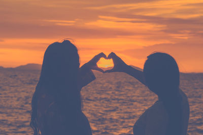 Silhouette friends making heart shaped while standing by sea against orange sky