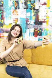 Portrait of young woman sitting on sofa at home and taking a selfie with avocado