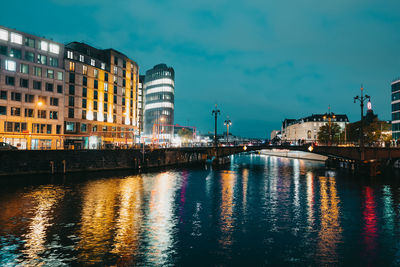 Illuminated buildings by river against sky at dusk