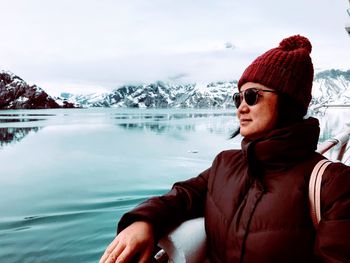 Woman wearing sunglasses by sea and snowcapped mountains against cloudy sky