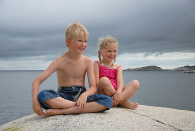 Smiling siblings sitting on rock formation with sea in background against cloudy sky
