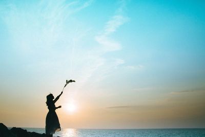 Woman standing with arms raised holding plant by sea against sky during sunset