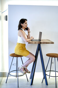 Female entrepreneur talking mobile phone while using netbook and working remotely at table in cafe