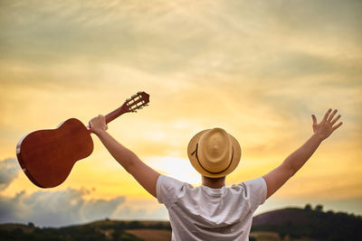 Rear view of man with arms raised standing against sky during sunset