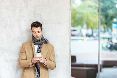 Young man using smart phone while standing against wall