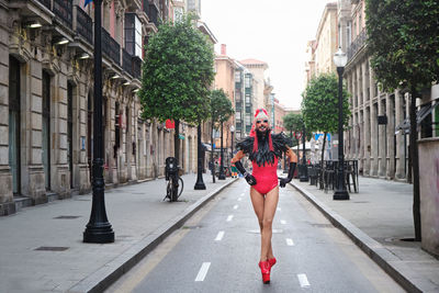 Full body of eccentric bearded male with makeup wearing red bodysuit with headdress and high heeled shoes strolling along asphalt road in city