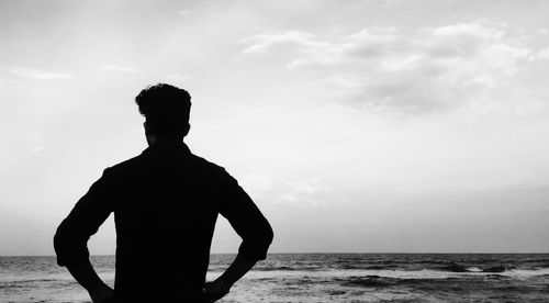 Rear view of silhouette man with hands on hip standing at beach against sky