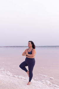 Serene curvy female in activewear standing in vrksasana on shore of pink lake and practicing yoga