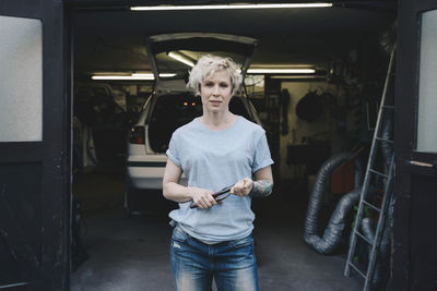 Portrait of female owner holding work tool while standing at entrance of auto repair shop