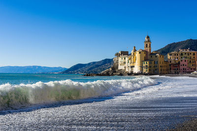 Sea waves rushing towards shore by buildings against clear blue sky