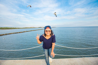 Portrait of smiling mid adult woman gesturing victory sign against sea and sky