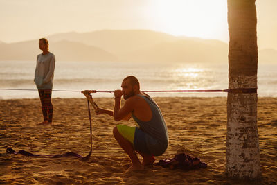 Side view of man holding rope while crouching on sand at beach against sky