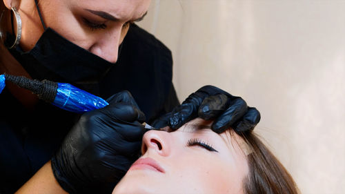 Close-up of beautician wearing mask coloring woman eyebrow in salon