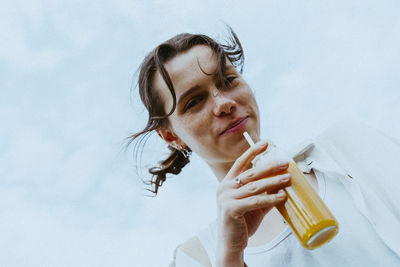 Low angle portrait of smiling young non-binary person holding juice bottle against sky