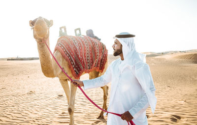 Man with camel standing on sand dunes at desert against sky