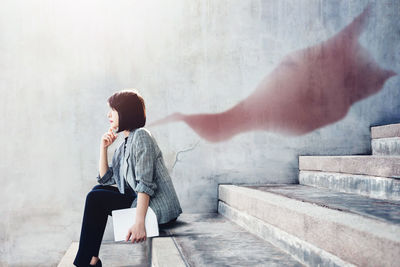 Side view of mature woman holding laptop while sitting on steps against wall