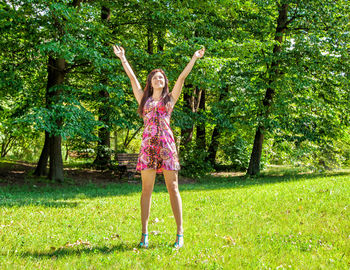 Full length of happy young woman with arms raised standing on grassy field in park