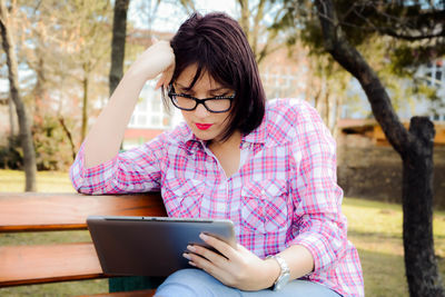 Young woman using digital tablet while sitting on bench at park